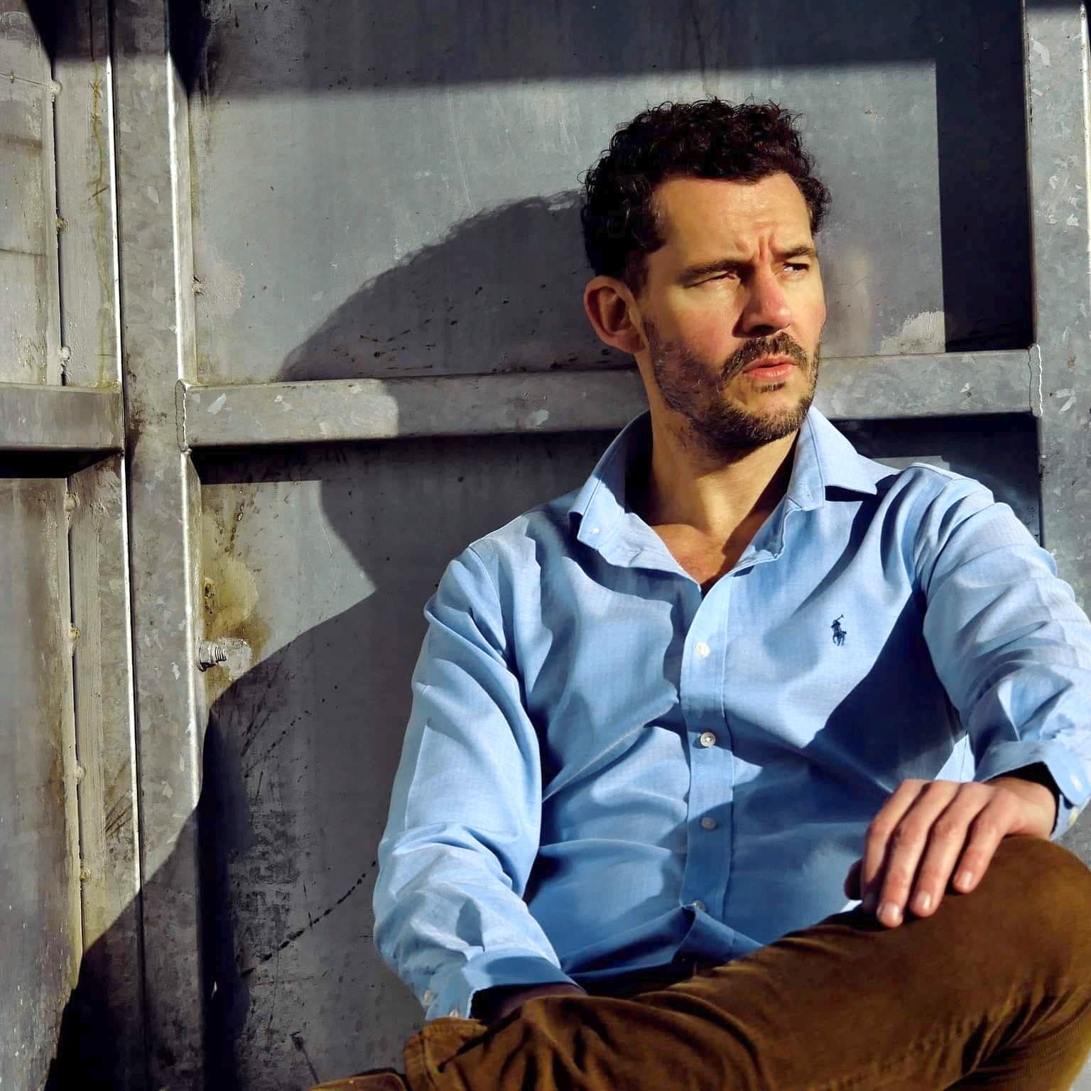 DJ Mark Loverush sitting down, looking pensively to the right. He is wearing a blue shirt and is leaning against a steel door.